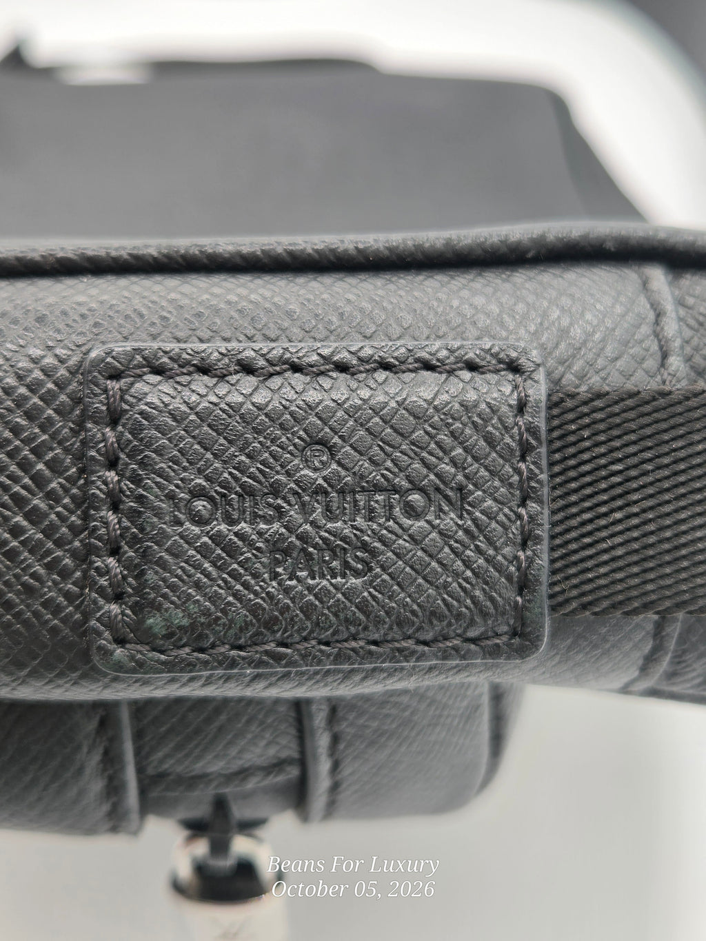 Close-up of a Louis Vuitton handbag in dark gray textured canvas, showing the embossed brand patch on a leather tab and part of the strap.