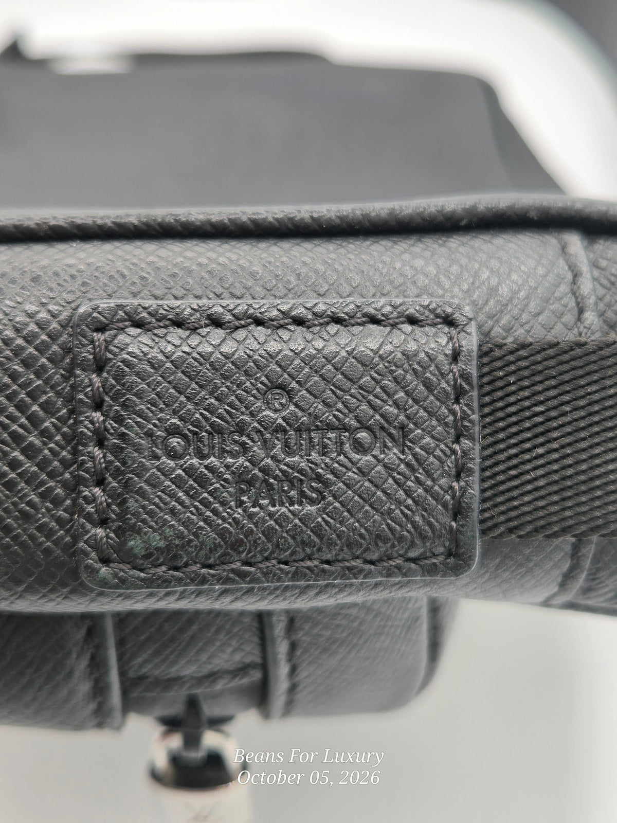Close-up of a Louis Vuitton handbag in dark gray textured canvas, showing the embossed brand patch on a leather tab and part of the strap.