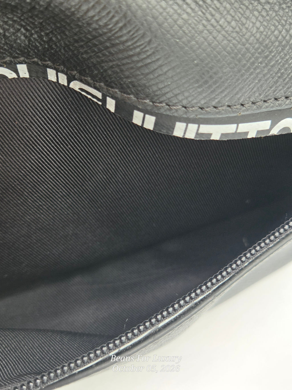 Close-up of the interior of a black textured leather pouch or clutch, showing a black fabric lining with a white printed logo and a zipper edge.