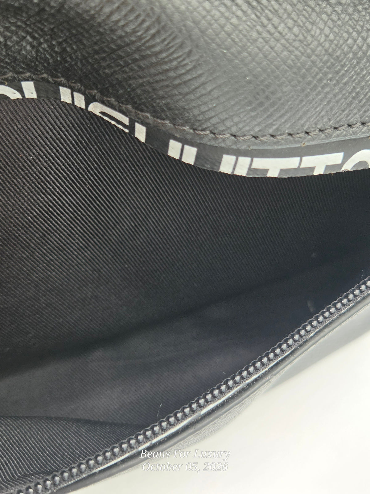 Close-up of the interior of a black textured leather pouch or clutch, showing a black fabric lining with a white printed logo and a zipper edge.