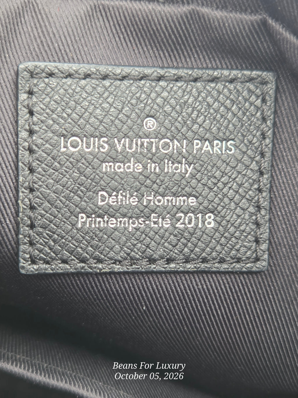 Close-up of a Louis Vuitton Paris leather interior label reading 'made in Italy', 'Défilé Homme', and 'Printemps-Eté 2018', stitched onto dark gray fabric.