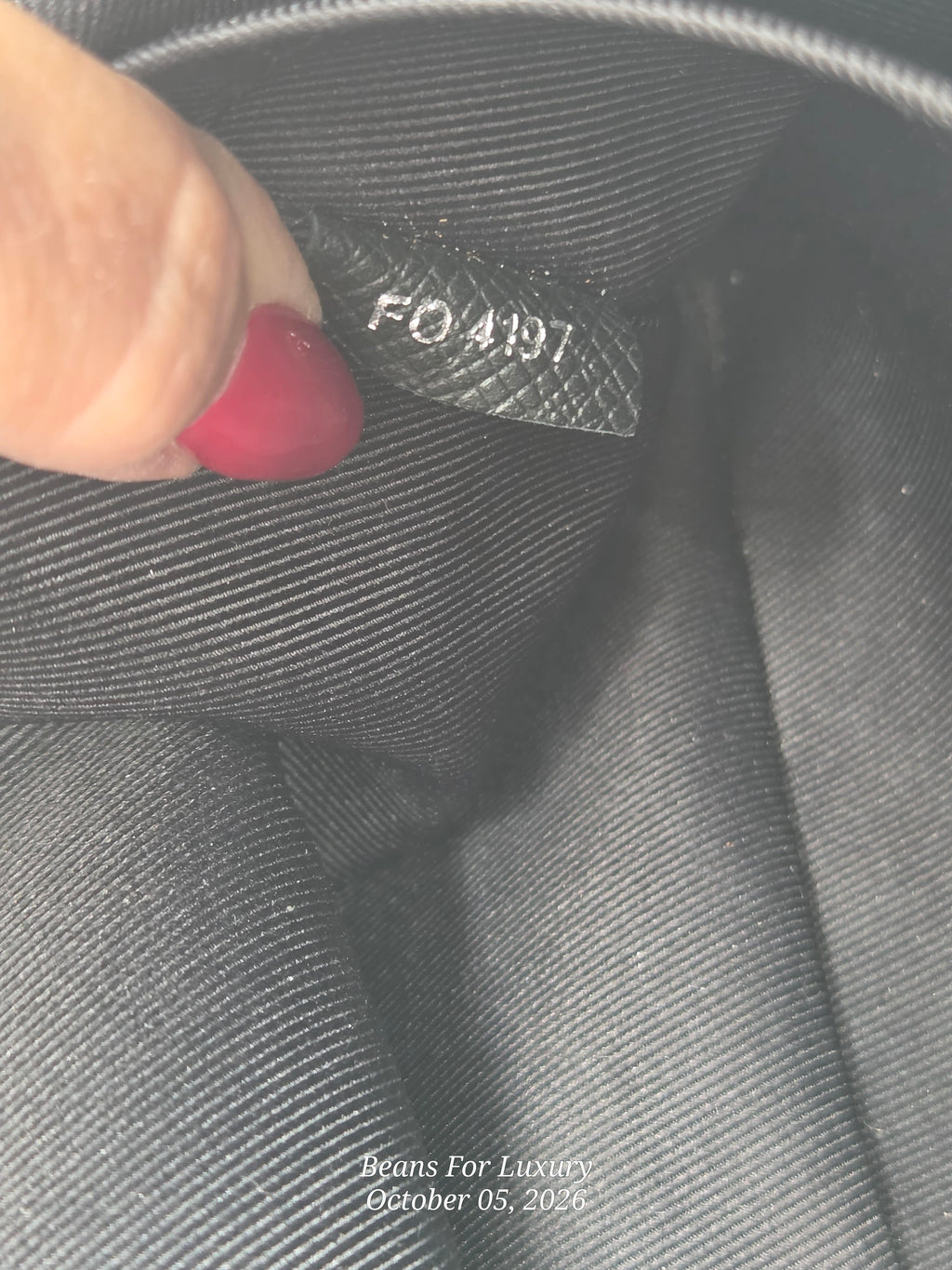 Close-up of a black fabric interior tag with embossed text reading 'FO4197', held by a finger with red nail polish against dark gray fabric.