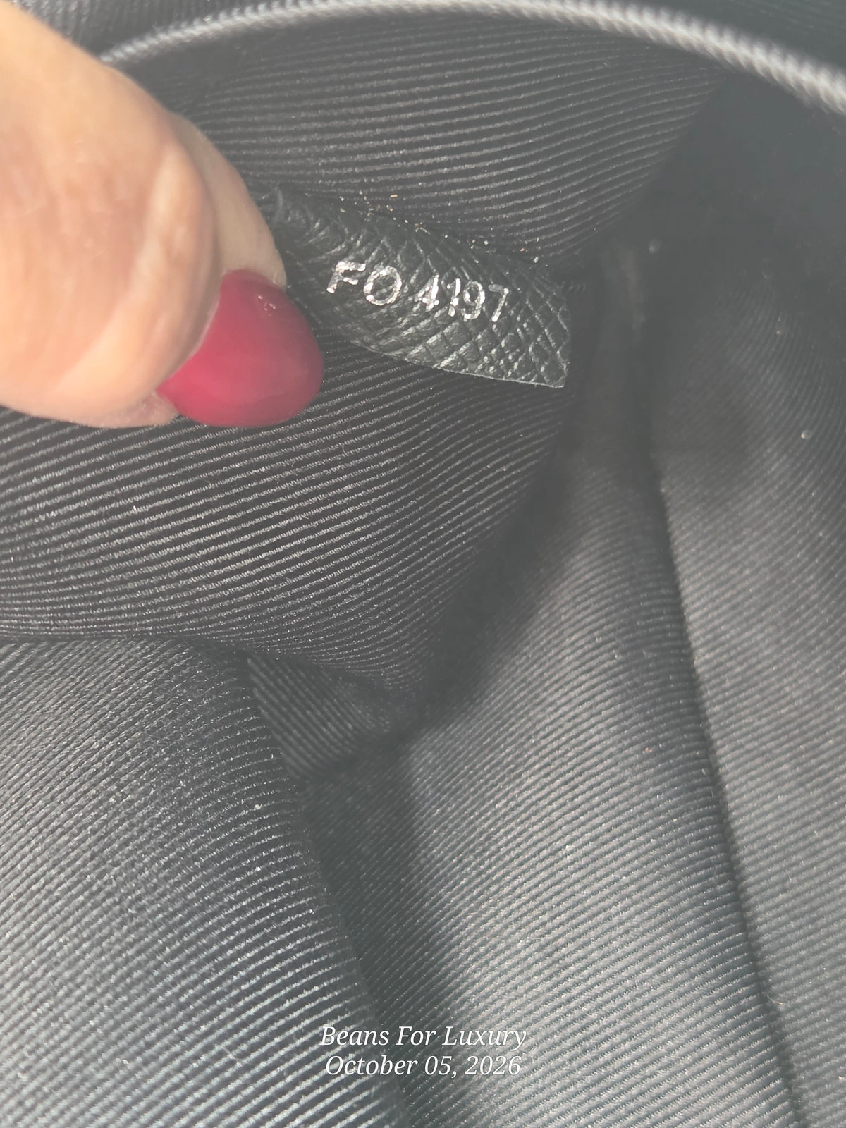 Close-up of a black fabric interior tag with embossed text reading 'FO4197', held by a finger with red nail polish against dark gray fabric.
