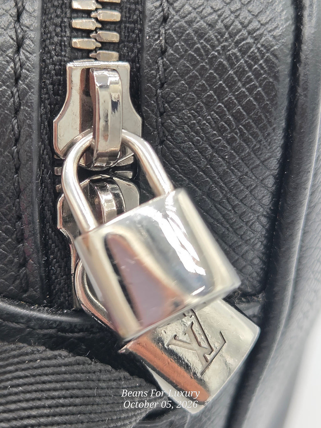 Close-up of a black textured leather bag with a silver padlock zipper pull featuring an embossed Louis Vuitton monogram, with a watermark in the bottom right corner.