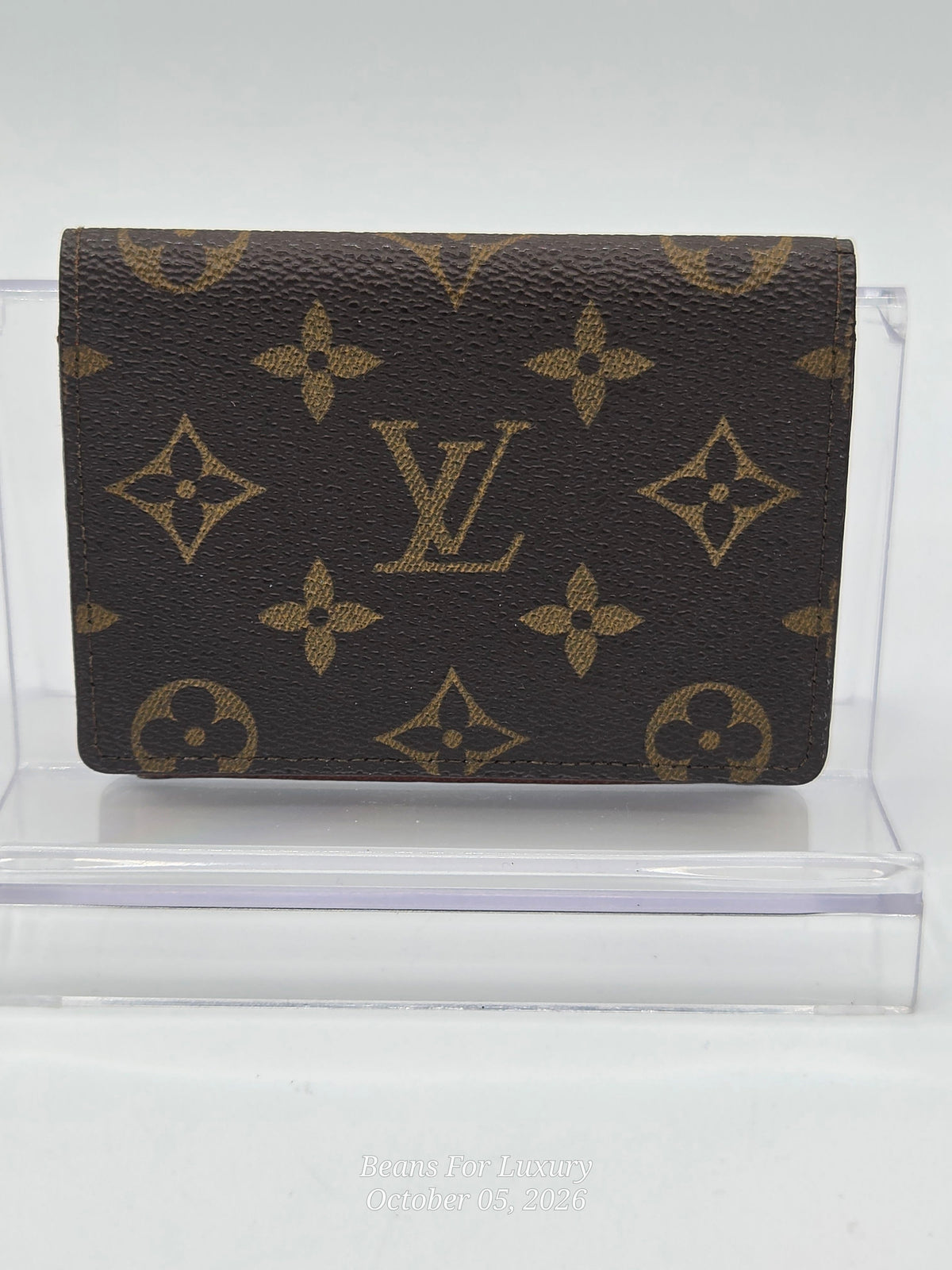 A Louis Vuitton monogram canvas wallet in dark brown with tan/gold printed logos and floral motifs, displayed on a clear acrylic stand against a light gray background.