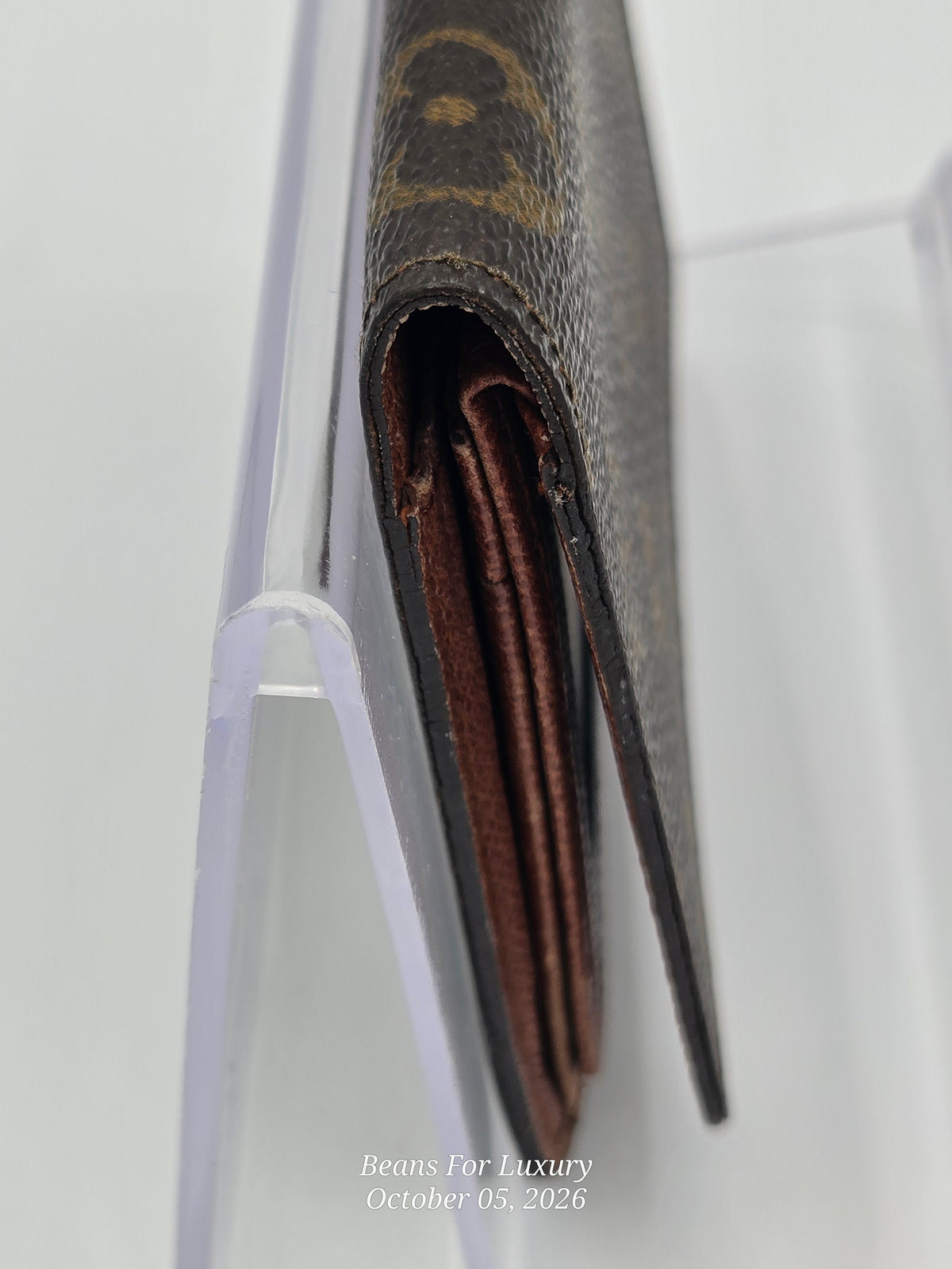 A close-up of a brown leather wallet with a dark monogram pattern and visible wear on the edges, displayed on a clear acrylic stand against a white background.