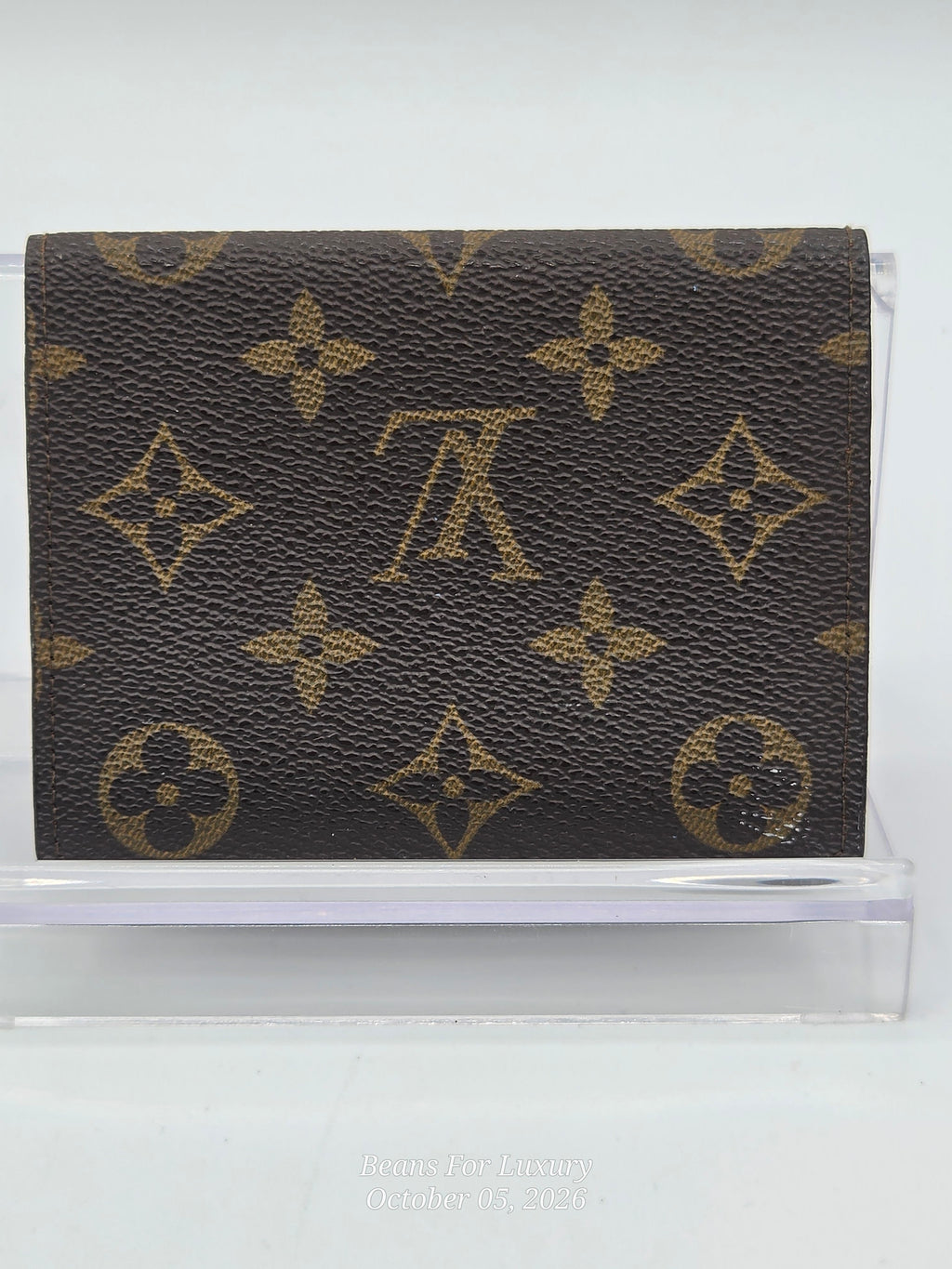 A Louis Vuitton monogram card holder wallet in dark brown coated canvas with the classic tan/gold LV monogram pattern, displayed on a clear acrylic stand.