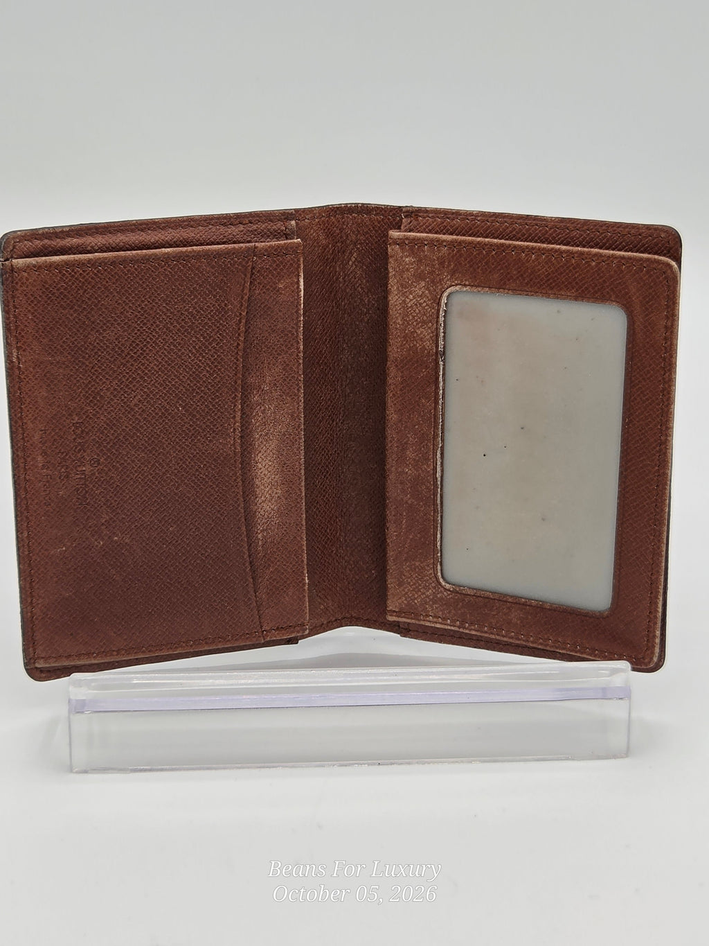 A brown textured leather bifold wallet shown open, displaying its interior card slots and a clear plastic ID window, resting on a clear acrylic display block against a light gray background.
