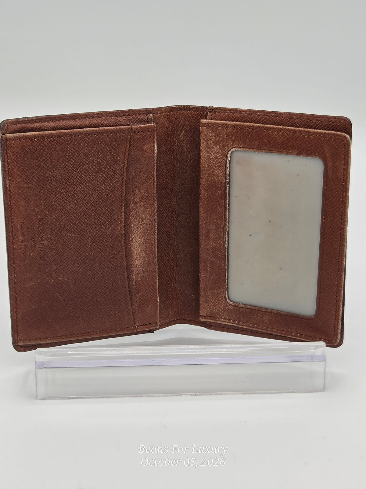 A brown textured leather bifold wallet shown open, displaying its interior card slots and a clear plastic ID window, resting on a clear acrylic display block against a light gray background.
