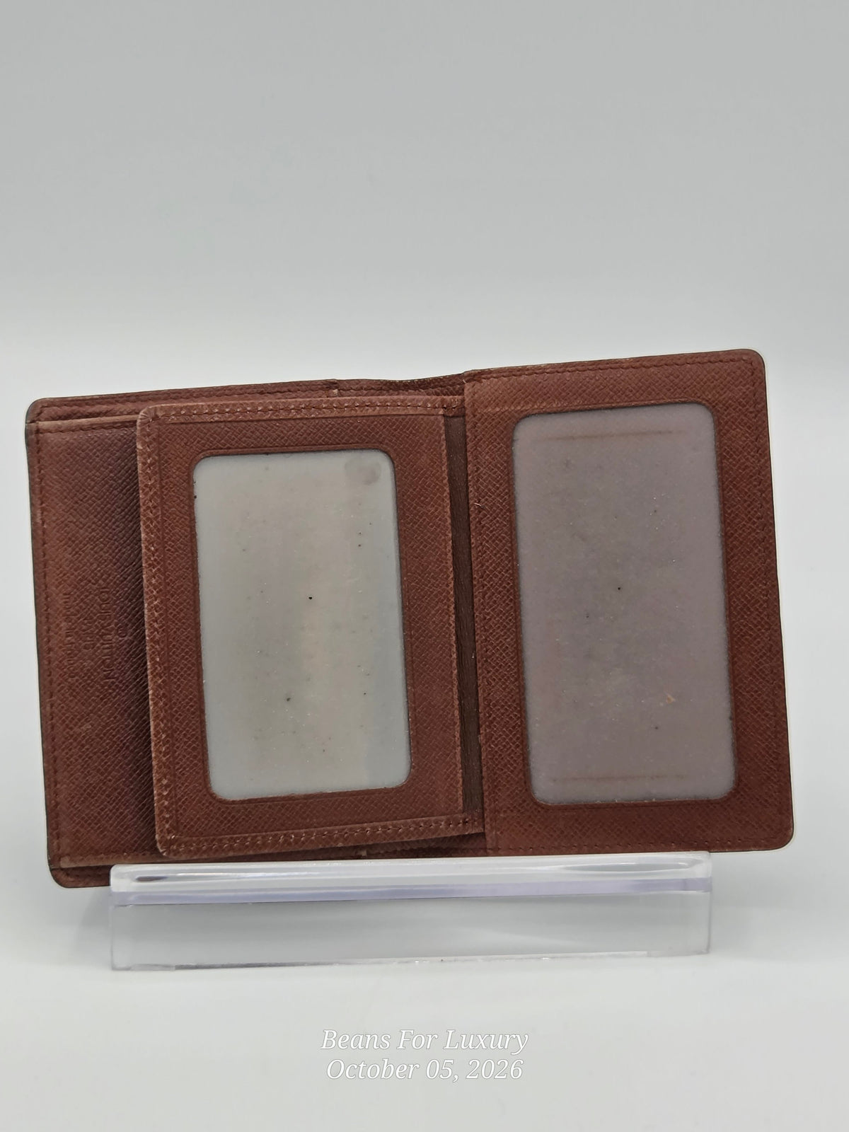 A brown textured leather bi-fold wallet shown open, displaying two clear ID slots, resting on a clear acrylic display stand against a light gray background.