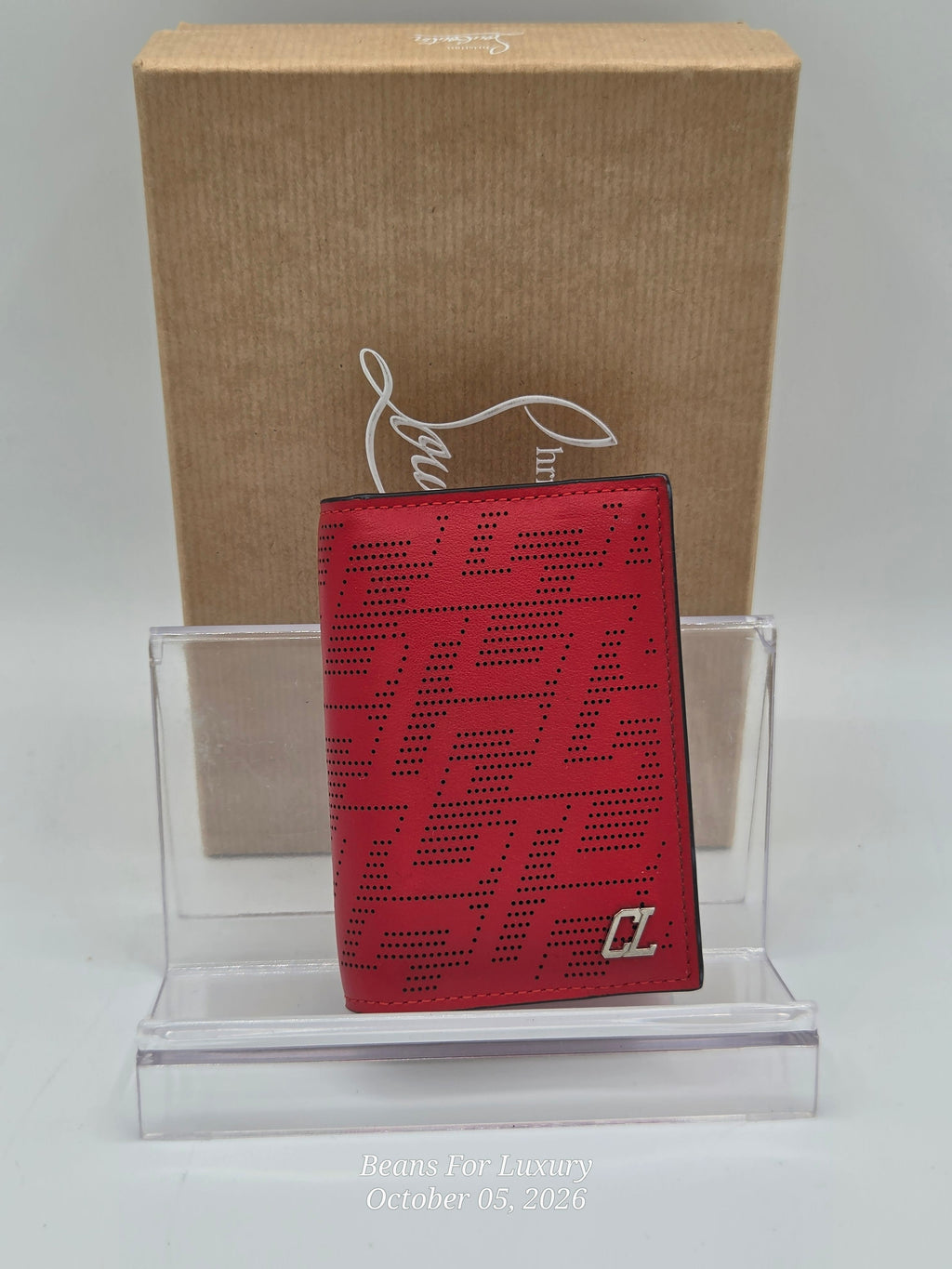 A red leather wallet with a perforated geometric pattern and a silver-toned 'L' logo, displayed on a clear acrylic stand in front of a brown cardboard box with a white logo.