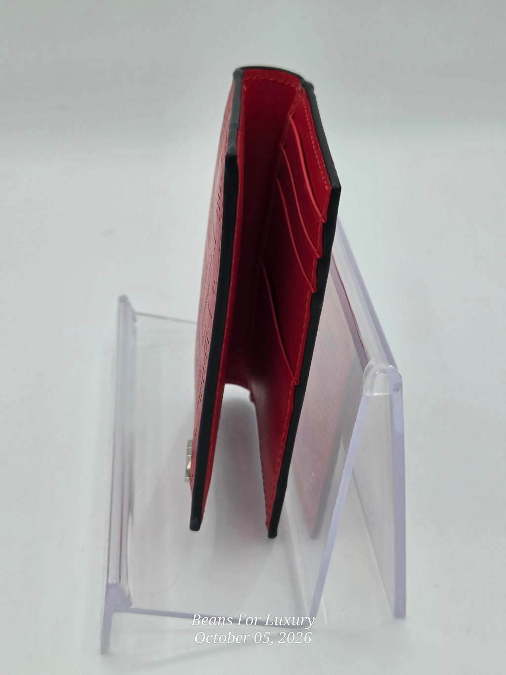 A slim red leather card holder wallet with black trim, displayed standing upright on a clear acrylic stand against a white background.