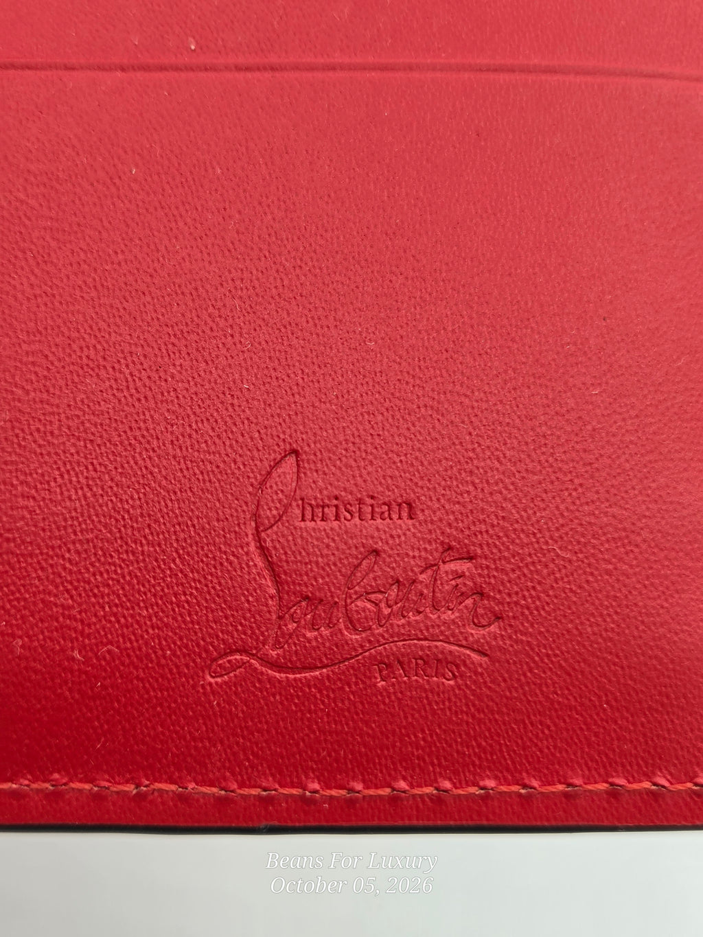 Close-up of a red textured leather item with debossed Christian Louboutin PARIS branding and matching red stitching along the bottom edge.