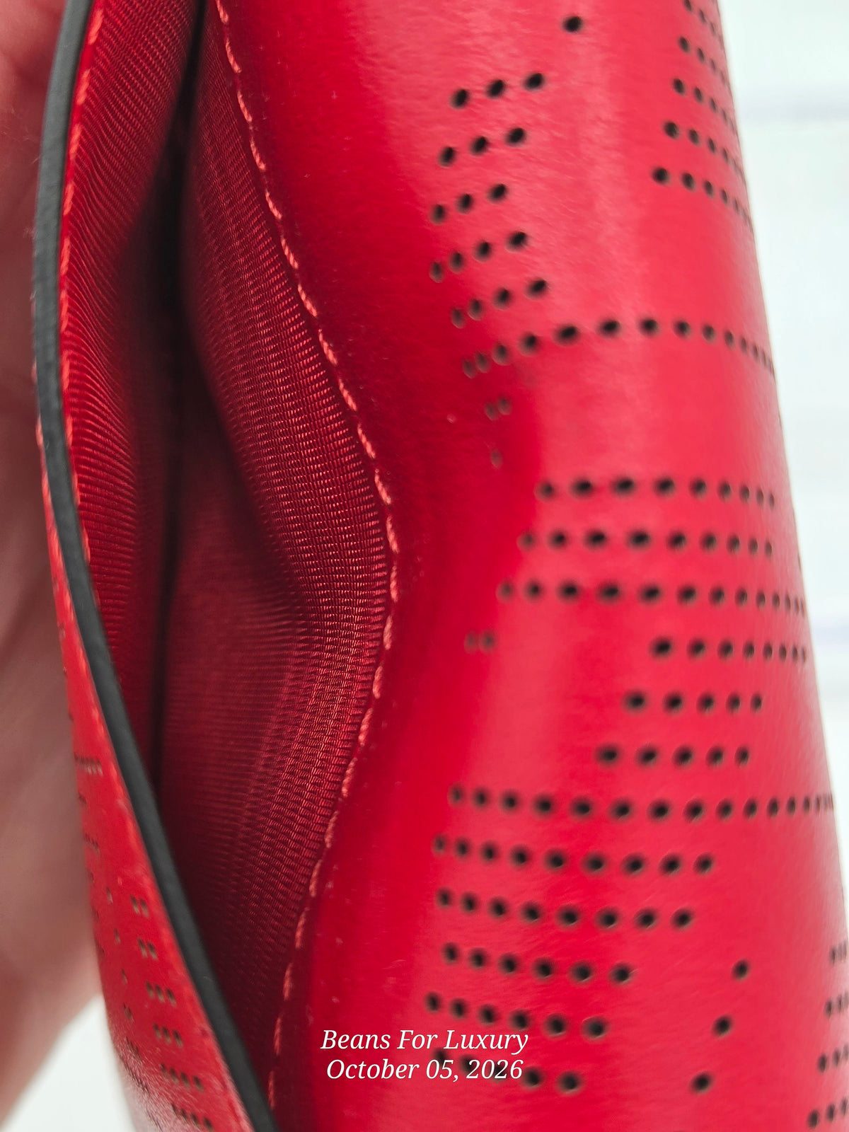 Close-up of a bright red leather item with perforated holes and visible interior stitching, held against a light background. Small white text at the bottom reads 'Beans For Luxury October 05, 2026'.