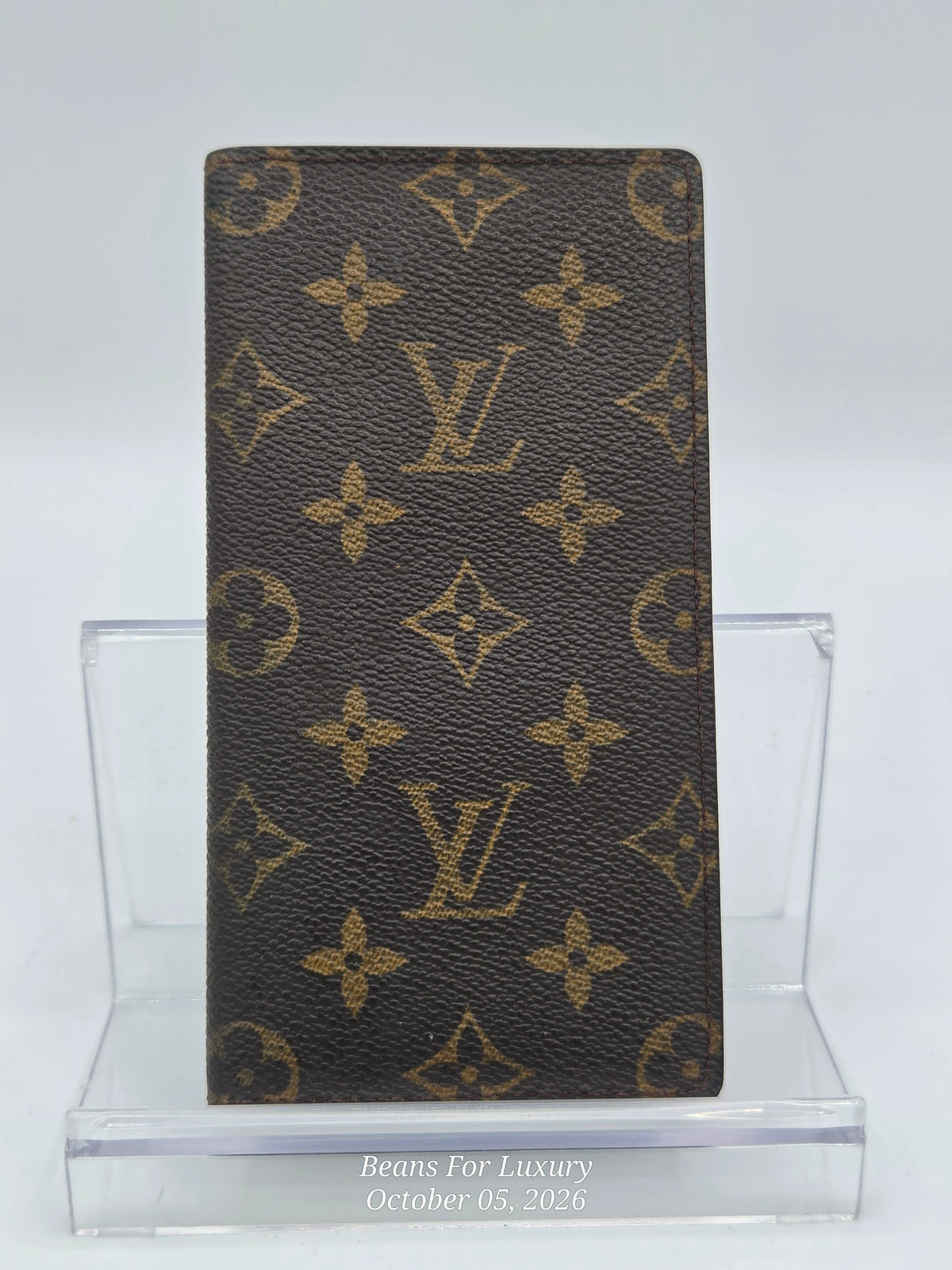 A Louis Vuitton monogram card holder or wallet made of dark brown coated canvas with the classic tan/gold LV monogram pattern, displayed on a clear acrylic stand.