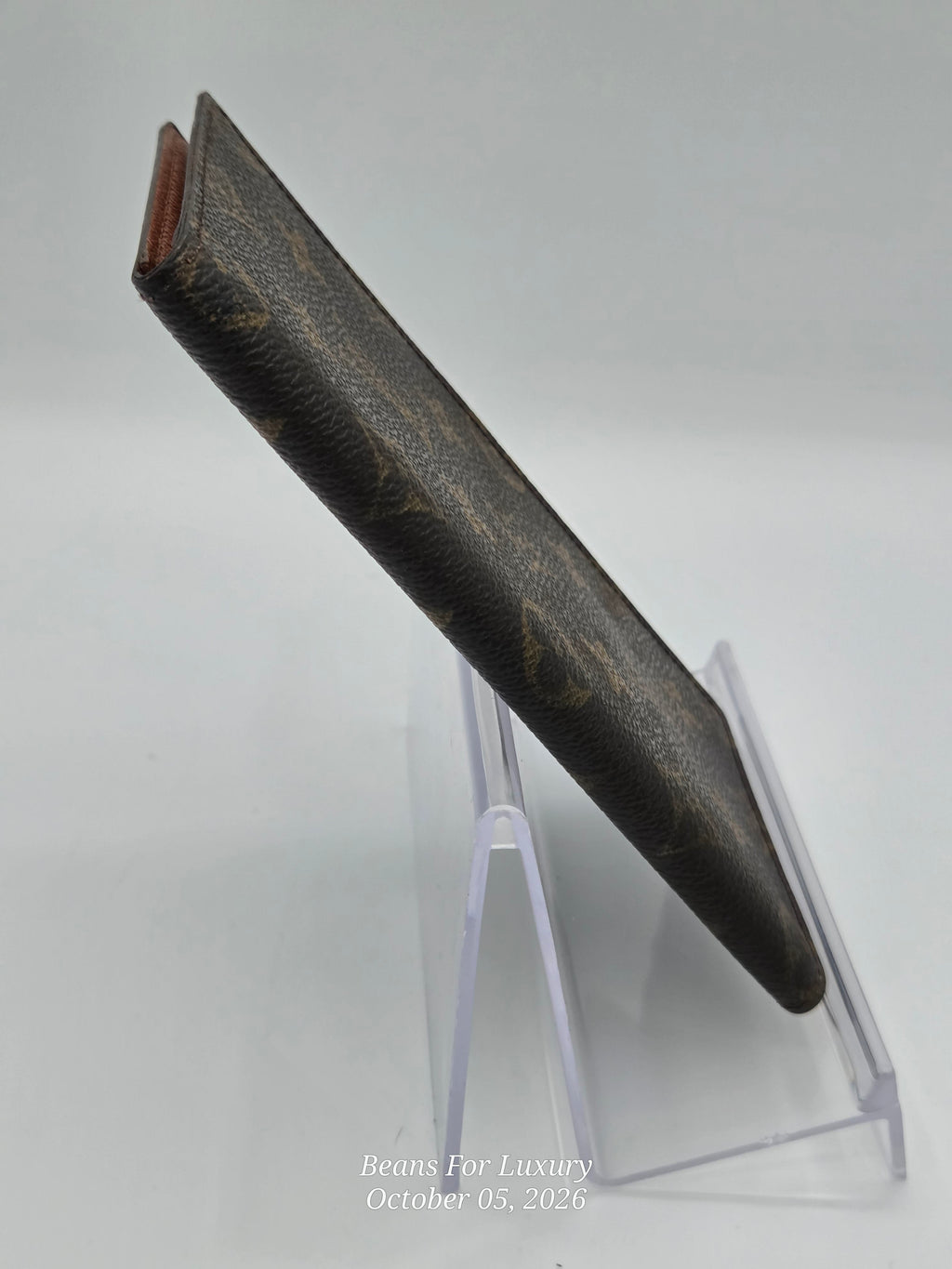 A dark brown Louis Vuitton monogram canvas sleeve or wallet standing upright on a clear acrylic display stand against a light gray background.