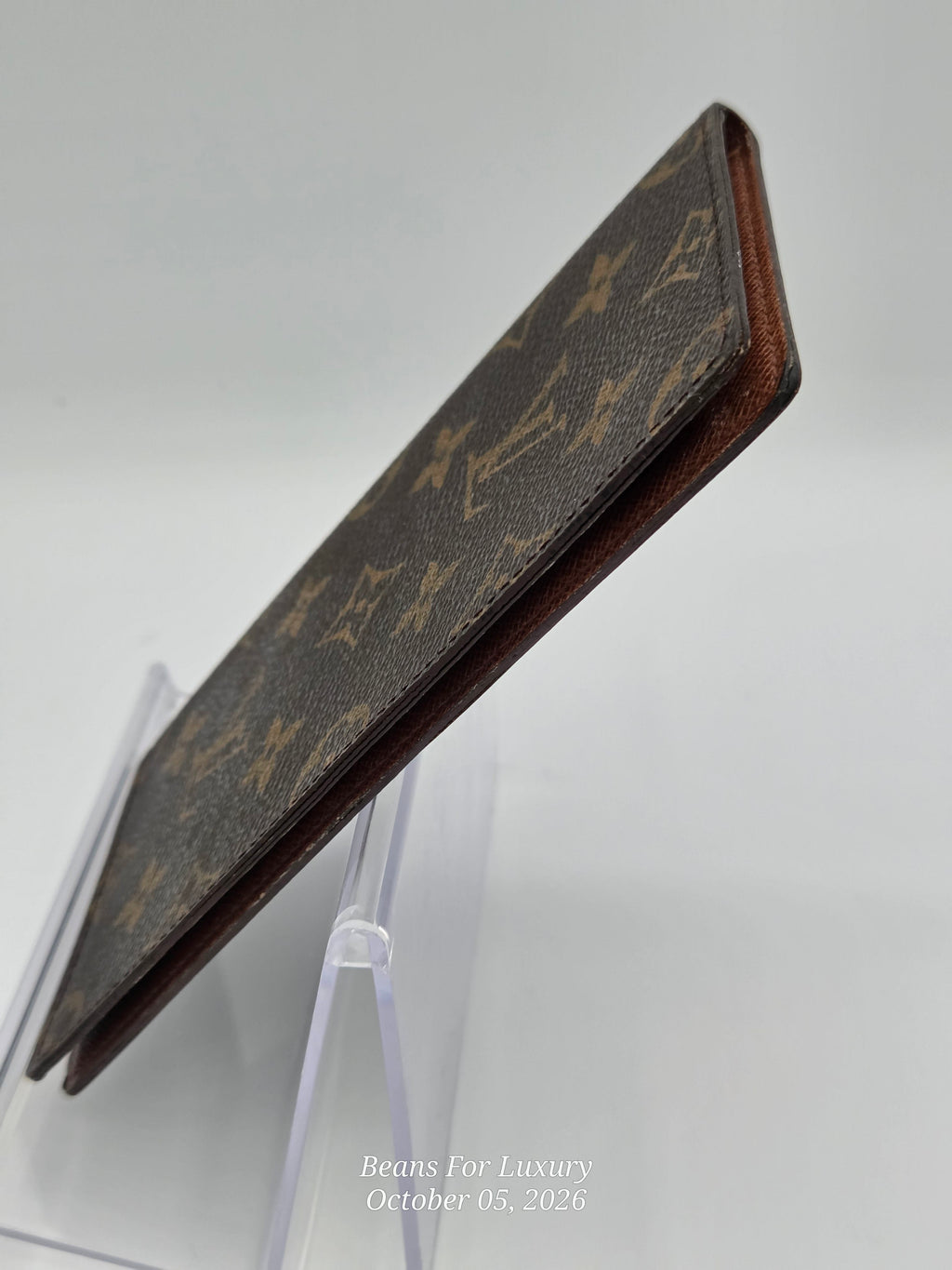 A Louis Vuitton monogram canvas card holder or wallet with brown leather edges, displayed on a clear acrylic stand against a light gray background.
