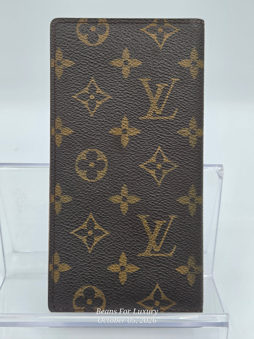 A Louis Vuitton monogram canvas checkbook holder or wallet in dark brown with tan monogram motifs, displayed upright on a clear acrylic stand against a light gray background.