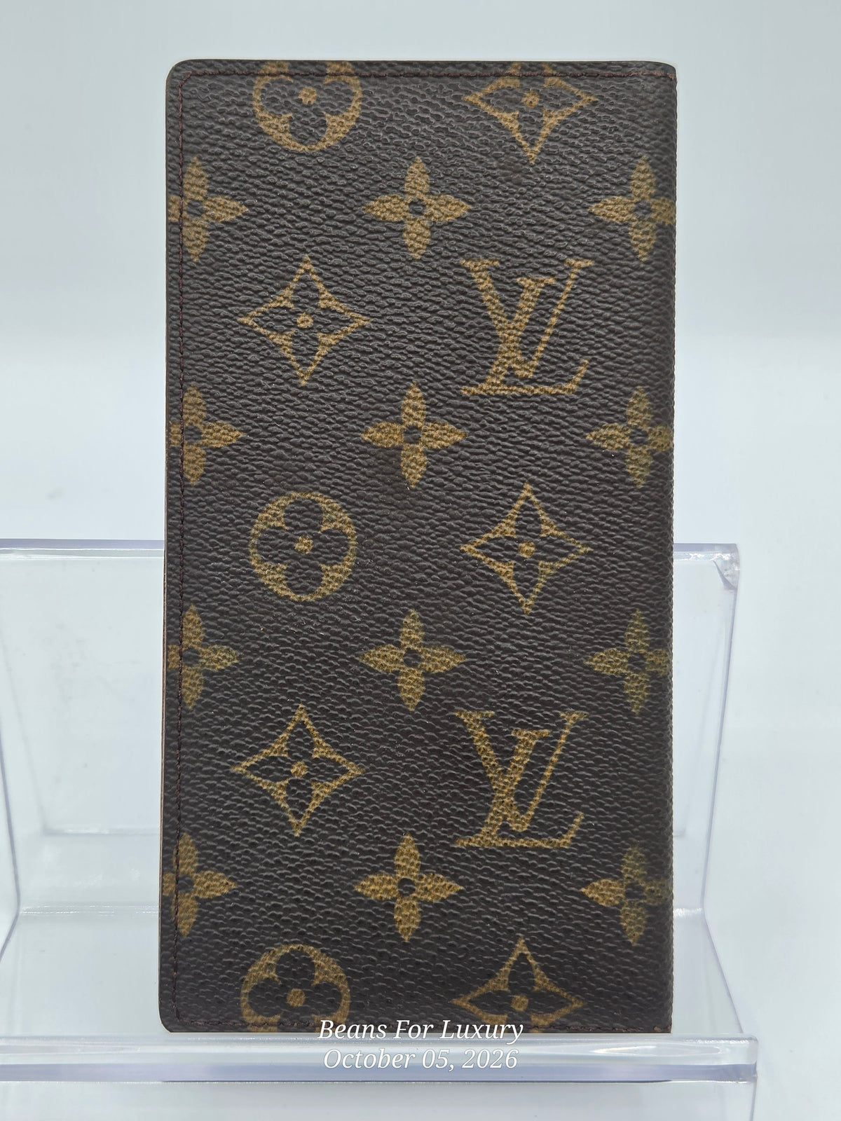 A Louis Vuitton monogram canvas checkbook holder or wallet in dark brown with tan monogram motifs, displayed upright on a clear acrylic stand against a light gray background.