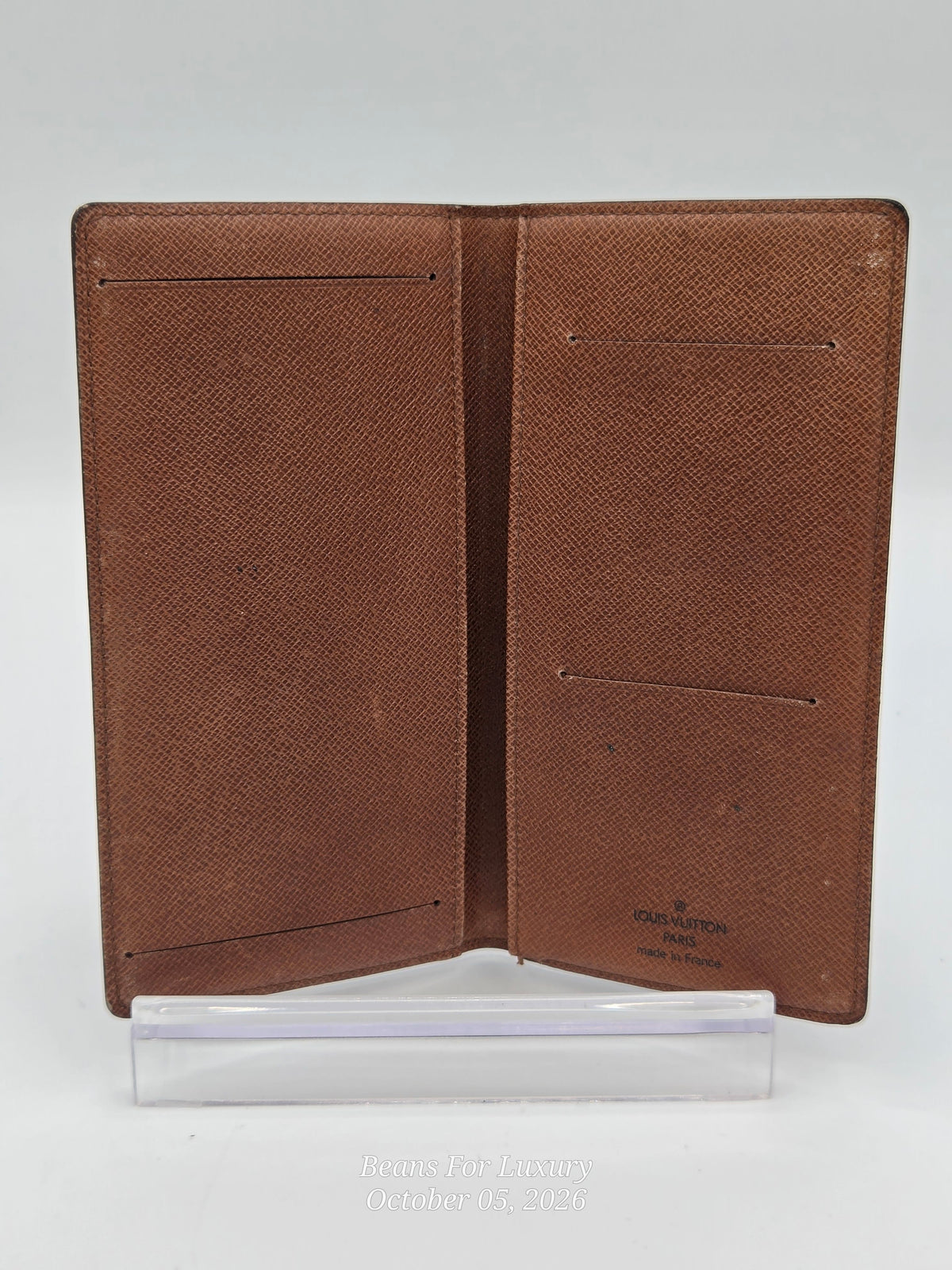 A brown textured leather passport cover shown open, displaying multiple interior card slots and pockets, standing on a clear plastic display block against a light gray background.
