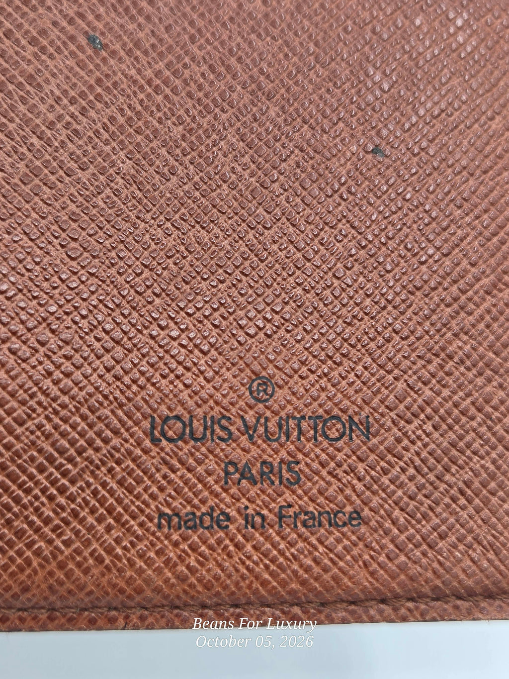 Close-up of a Louis Vuitton textured canvas material in a warm reddish-brown tone, with dark printed text reading 'LOUIS VUITTON PARIS made in France'.