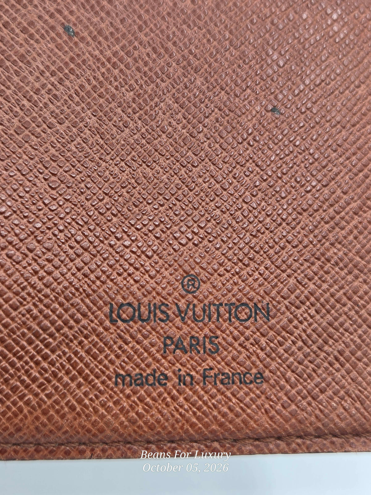 Close-up of a Louis Vuitton textured canvas material in a warm reddish-brown tone, with dark printed text reading 'LOUIS VUITTON PARIS made in France'.