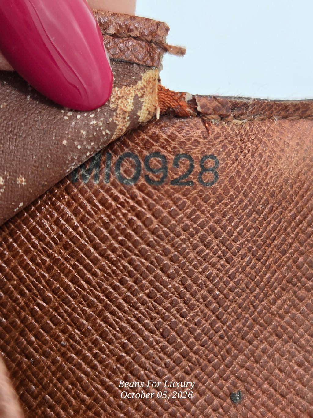 Close-up of a brown textured leather item with a peeling corner revealing a lighter inner layer, a pink manicured fingernail touching the edge, and a black stamped serial number 'M10928'.