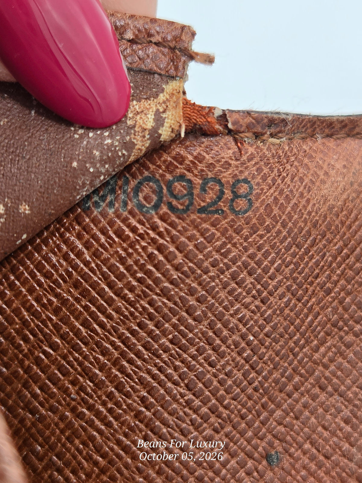 Close-up of a brown textured leather item with a peeling corner revealing a lighter inner layer, a pink manicured fingernail touching the edge, and a black stamped serial number 'M10928'.