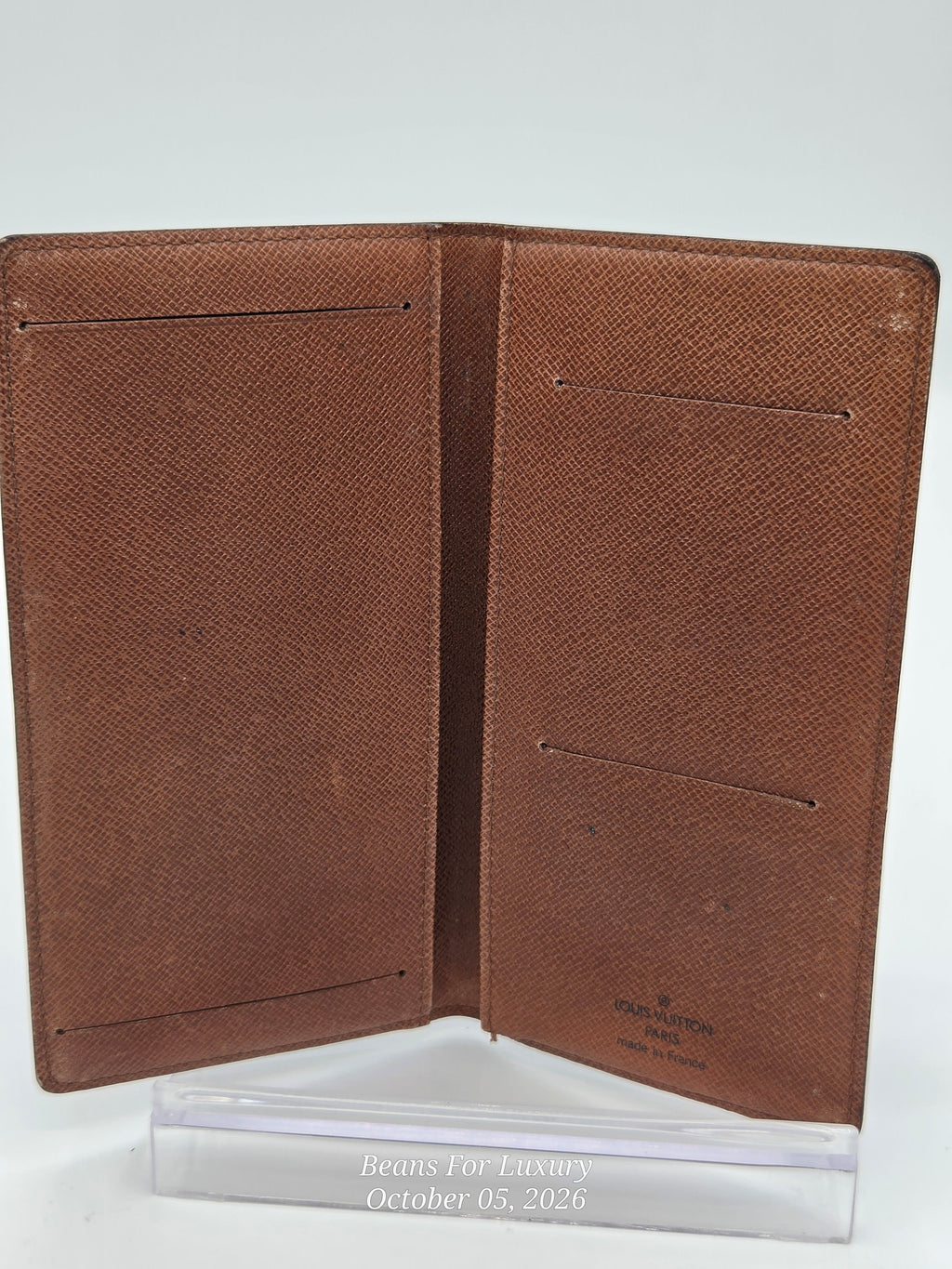 A brown textured leather passport cover or document holder shown open, photographed against a light gray background with a clear acrylic stand at the bottom. A small Louis Vuitton mark is visible near the lower right.