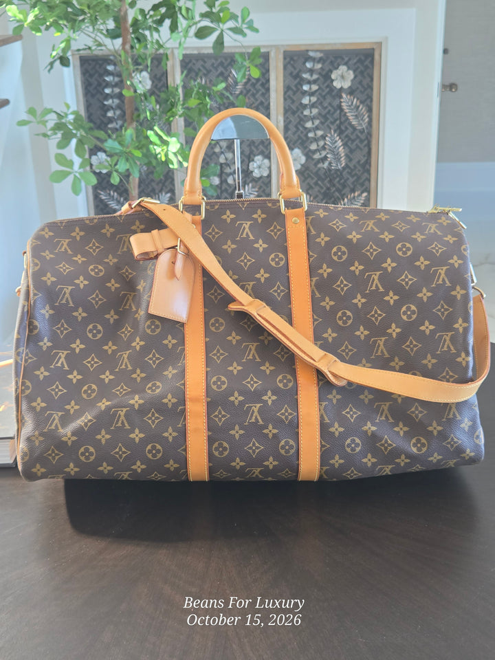 A Louis Vuitton monogram duffel bag in brown coated canvas with tan leather handles, trim, and shoulder strap, displayed on a dark wooden table in an indoor setting.
