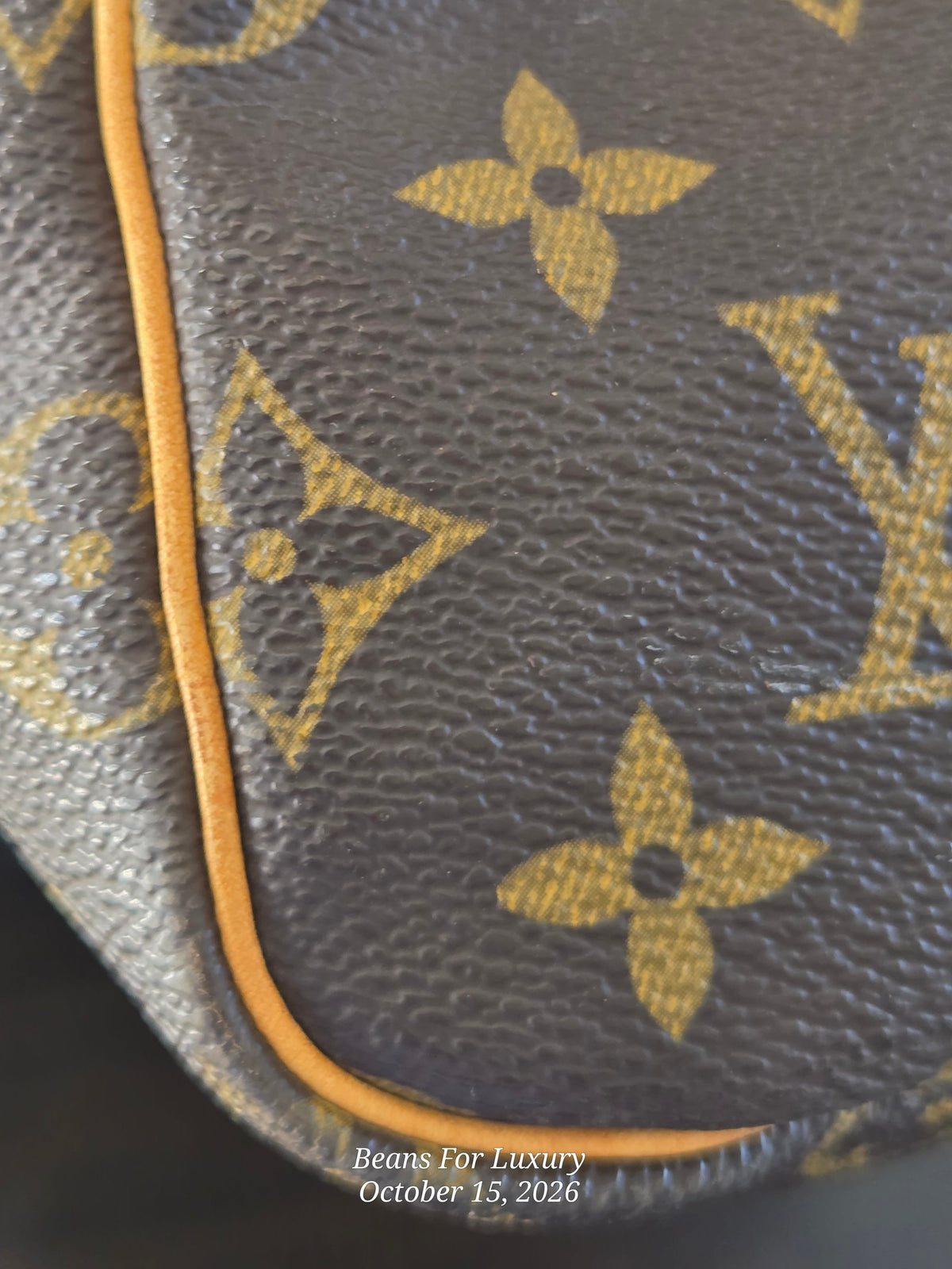 Close-up of a Louis Vuitton monogram coated canvas bag, showing the classic brown and gold LV pattern and a thin orange-yellow piping edge.