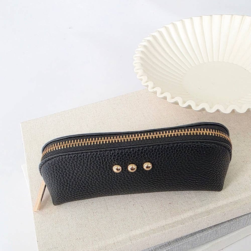 A black pebbled leather zippered pouch with three gold-tone rivets, resting on a beige textured book next to a white ceramic bowl.