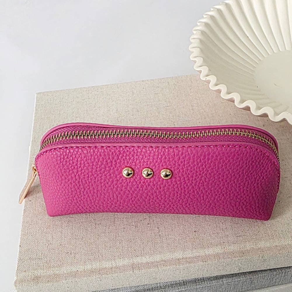 A bright pink textured leather pencil case with a gold zipper and three gold-tone rivets, resting on a beige linen book next to a white ceramic dish.