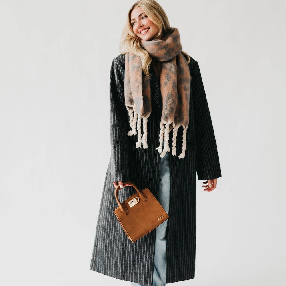 Woman wearing a long coat and patterned scarf, holding a brown handbag on a plain background