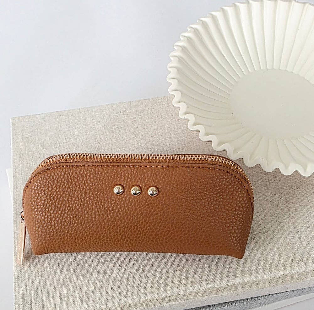 A brown pebbled faux-leather zippered pouch with three gold-tone rivets, resting on a beige linen book next to a white ceramic plate.
