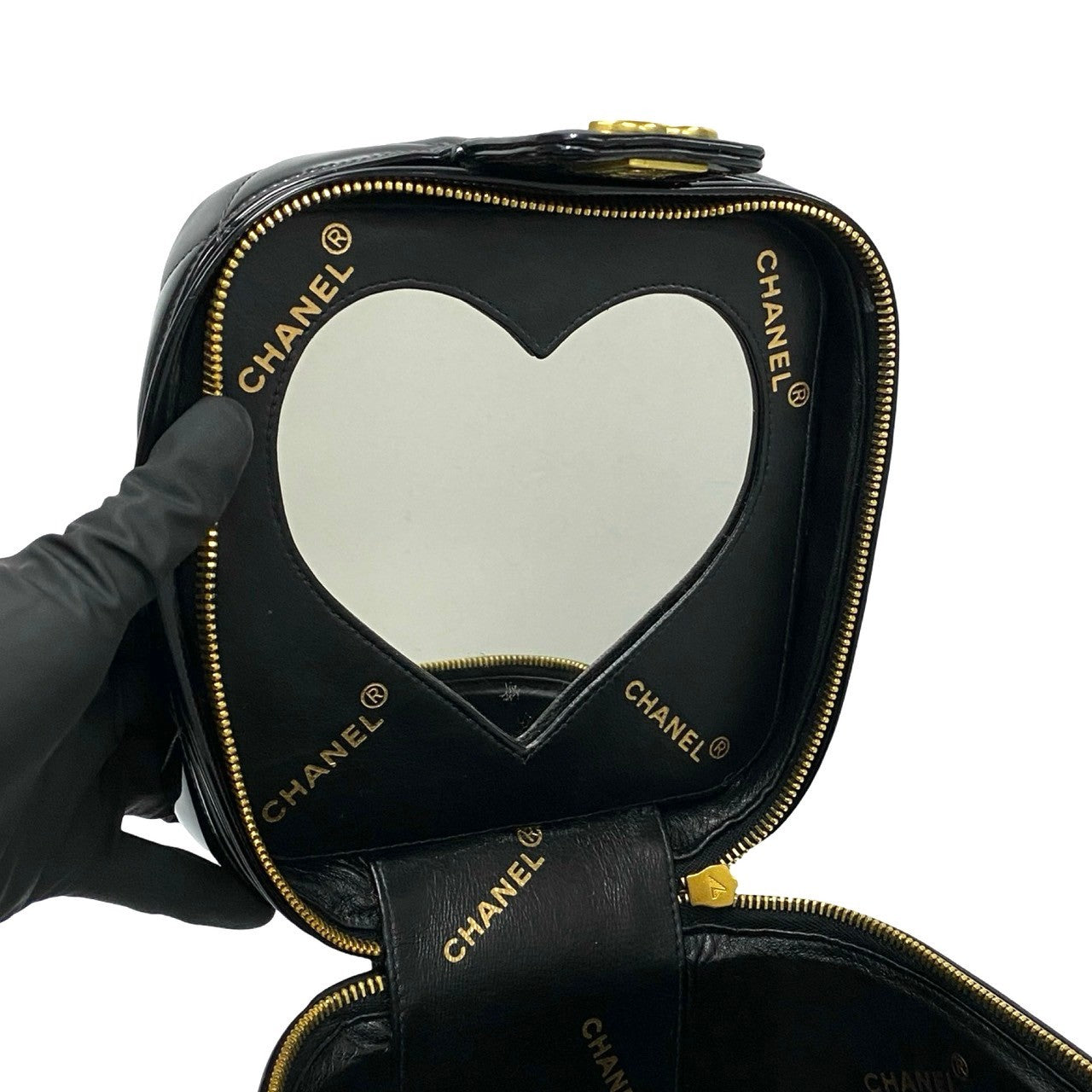 Black Chanel heart-shaped makeup case with gold accents held by a gloved hand on a white background.