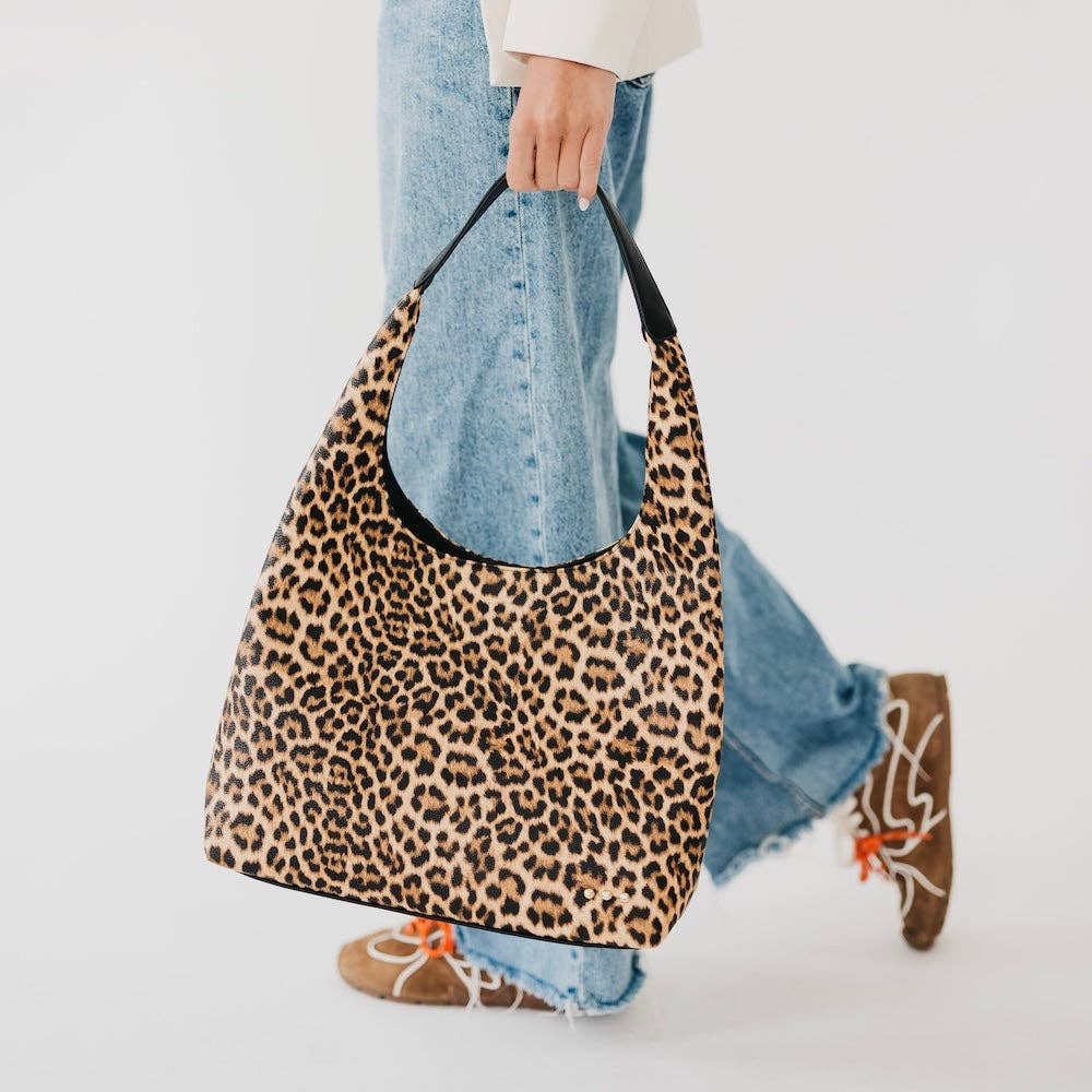 A person wearing blue jeans and brown lace-up shoes carries a large slouchy shoulder bag with a leopard print pattern against a clean white background.