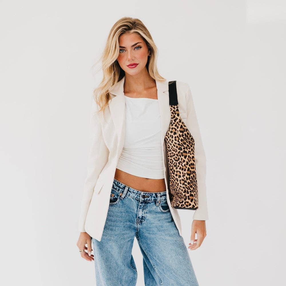 A woman models a leopard print shoulder tote bag with a black strap, styled over a white crop top and blazer with blue jeans against a plain white background.