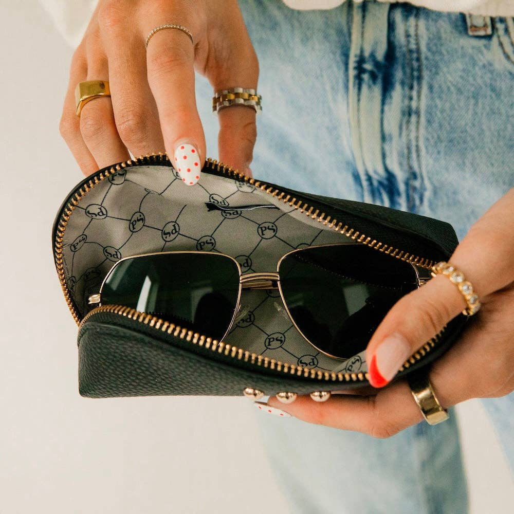A close-up of a person holding an open black pebbled leather sunglasses case with gold-tone hardware. The case has a repeating interlocking G pattern on the interior lining and holds dark aviator-style sunglasses.