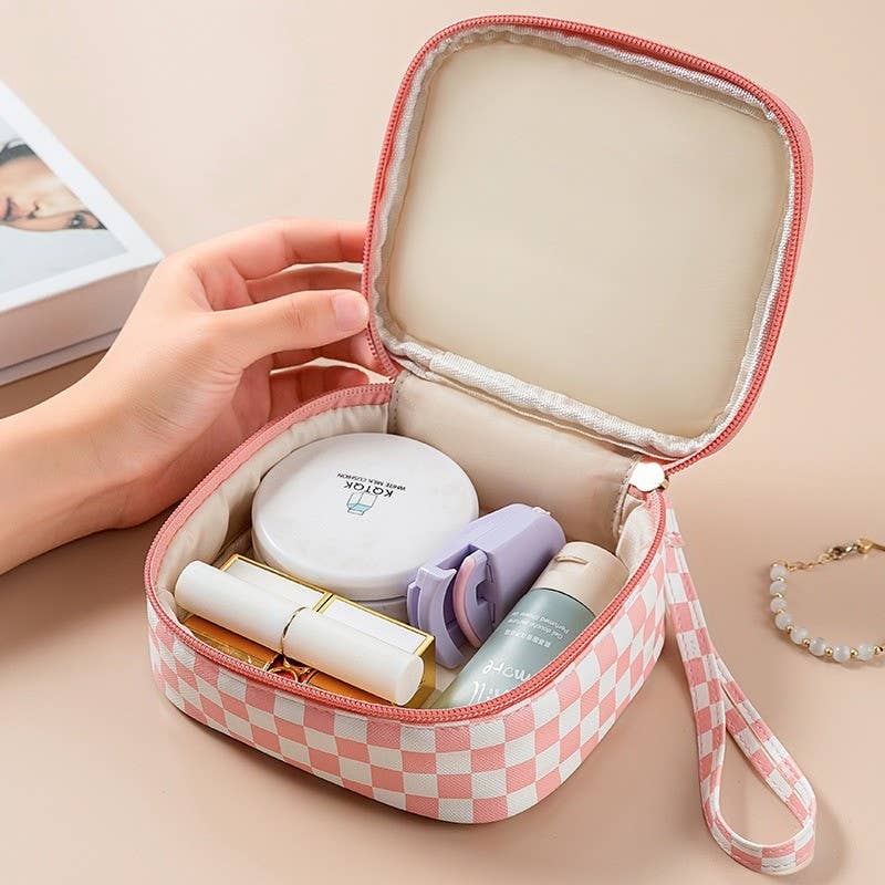 A pink and white checkered cosmetic bag with a handle, shown open with various makeup items inside. A hand is holding the bag, and a pearl bracelet and a book are visible in the background on a beige surface.