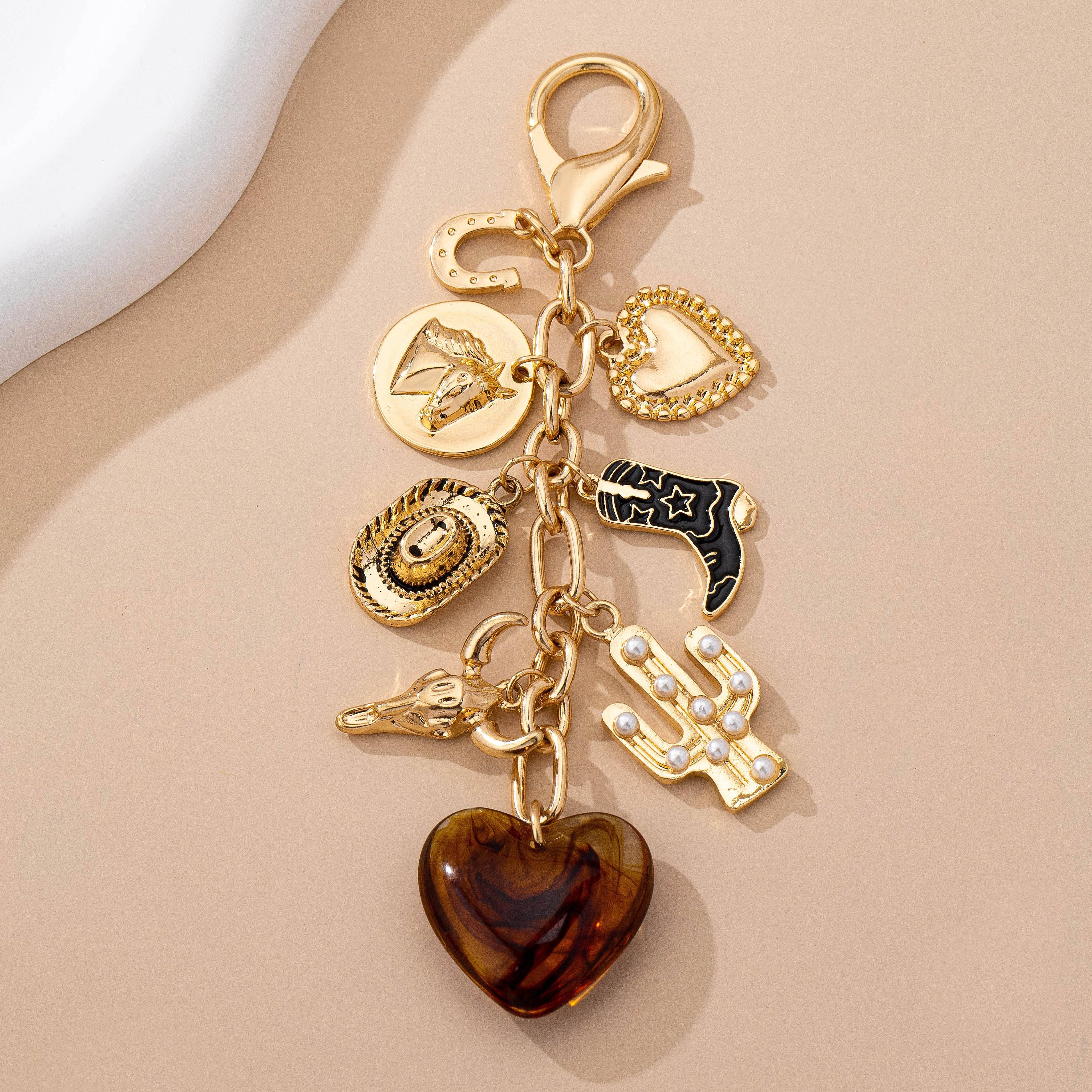 A gold-tone western-themed charm keychain with a lobster clasp, featuring multiple dangling charms including a brown heart, cowboy boot, horseshoe, horseshoe-shaped medallion, heart medallion, horseshoe silhouette, black-and-gold boot, horseshoe silhouette, cactus with pearl accents, and a brown translucent heart pendant, displayed on a beige background.