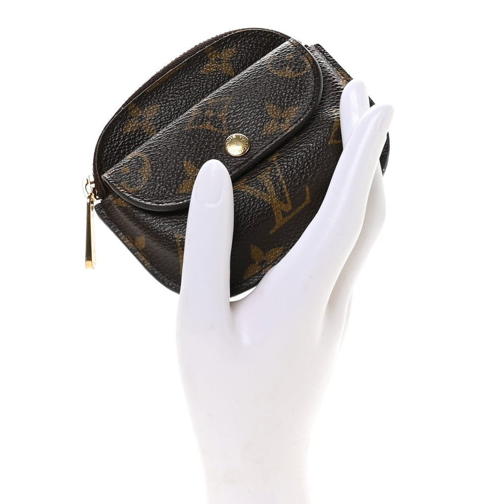 A Louis Vuitton monogram canvas coin purse held in a white mannequin hand against a plain white background.