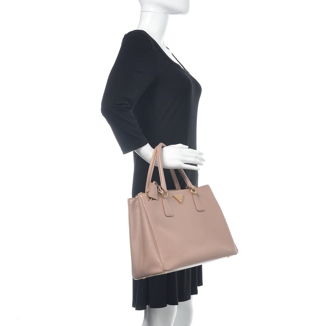 A mannequin wearing a black dress holds a large structured tote bag in a dusty blush pink color with gold-tone hardware and a small triangular logo charm, photographed against a plain white background.